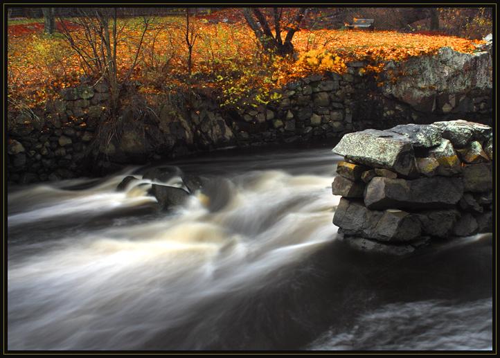 A section of the Ipswich River (Massachusetts, US) in the autumn. A slow shutter speed blurred the moving water. There is rushing water and bright yellow and green leaves.