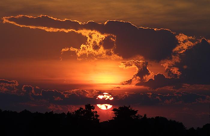 The sun setting behind some clouds. The edges of the clouds are brightly lit. Trees are silhouetted along the horizon. Taken in the salt marsh in Newbury, Massachusetts (US).