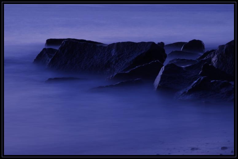 A long exposure photo of rocks on the beach. Taken after sunset. The long exposure allowed time for several waves to wash over the rocks, giving a misty look. Taken on Cape Cod, Massachusetts (US).